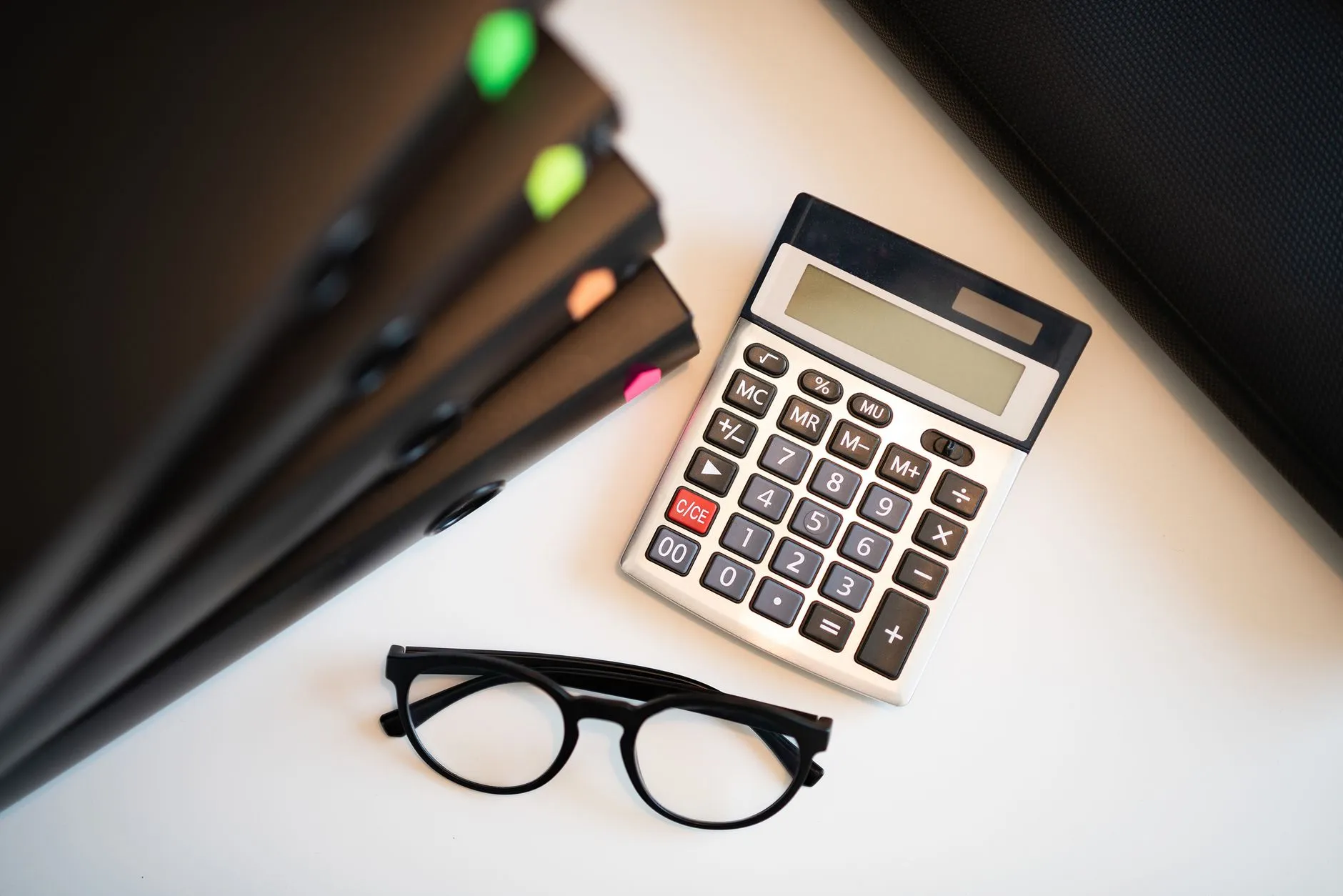 A calculator, reading glasses and a stack of document folders on a desk.